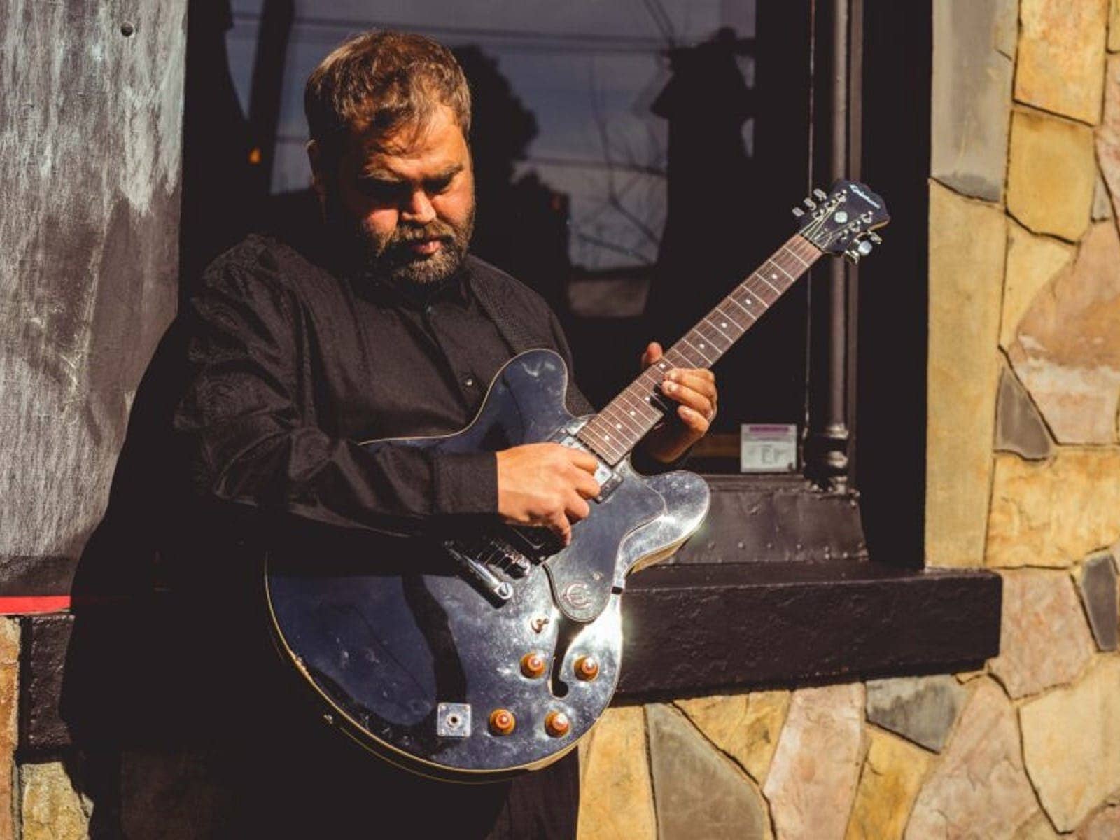 Robert K Champion is playing a black guitar leaning up against a window