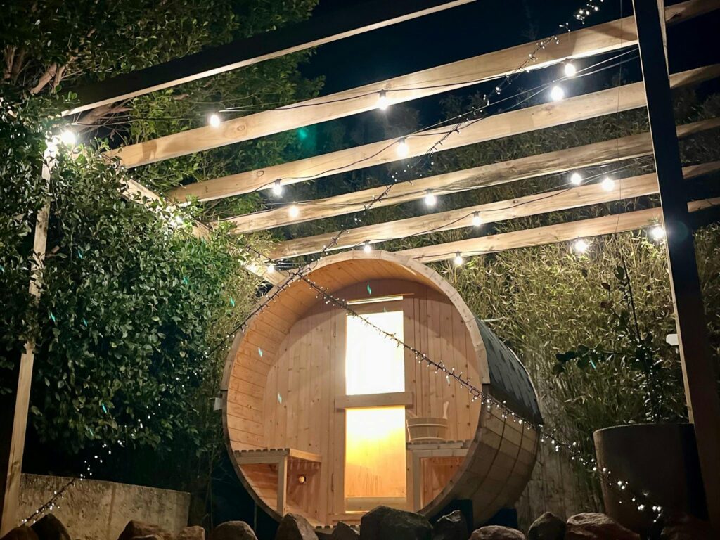 “Wooden barrel sauna with outdoor shower in a coastal garden setting, perfect for relaxation.”