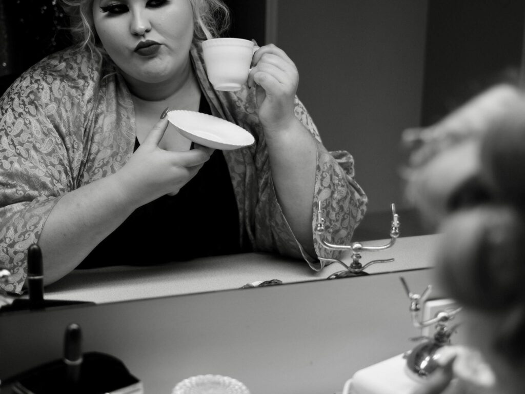 Charlee Watt with hair in curlers holding a coffee cup and gazing into a mirror, contemplating.