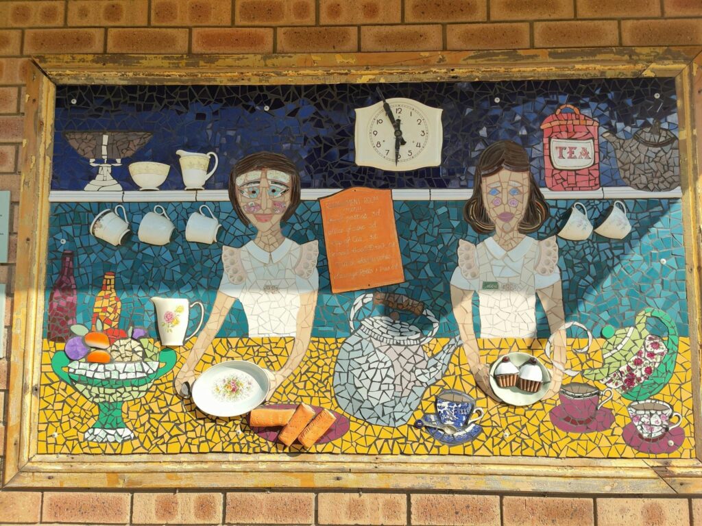 A colourful café-themed mosaic on the Cummins Mosaic Trail, showing two servers with teacups and cak