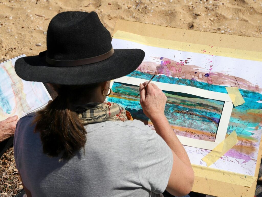 A woman in a wide brim hat is sitting and painting in watercolour while sitting on sand.